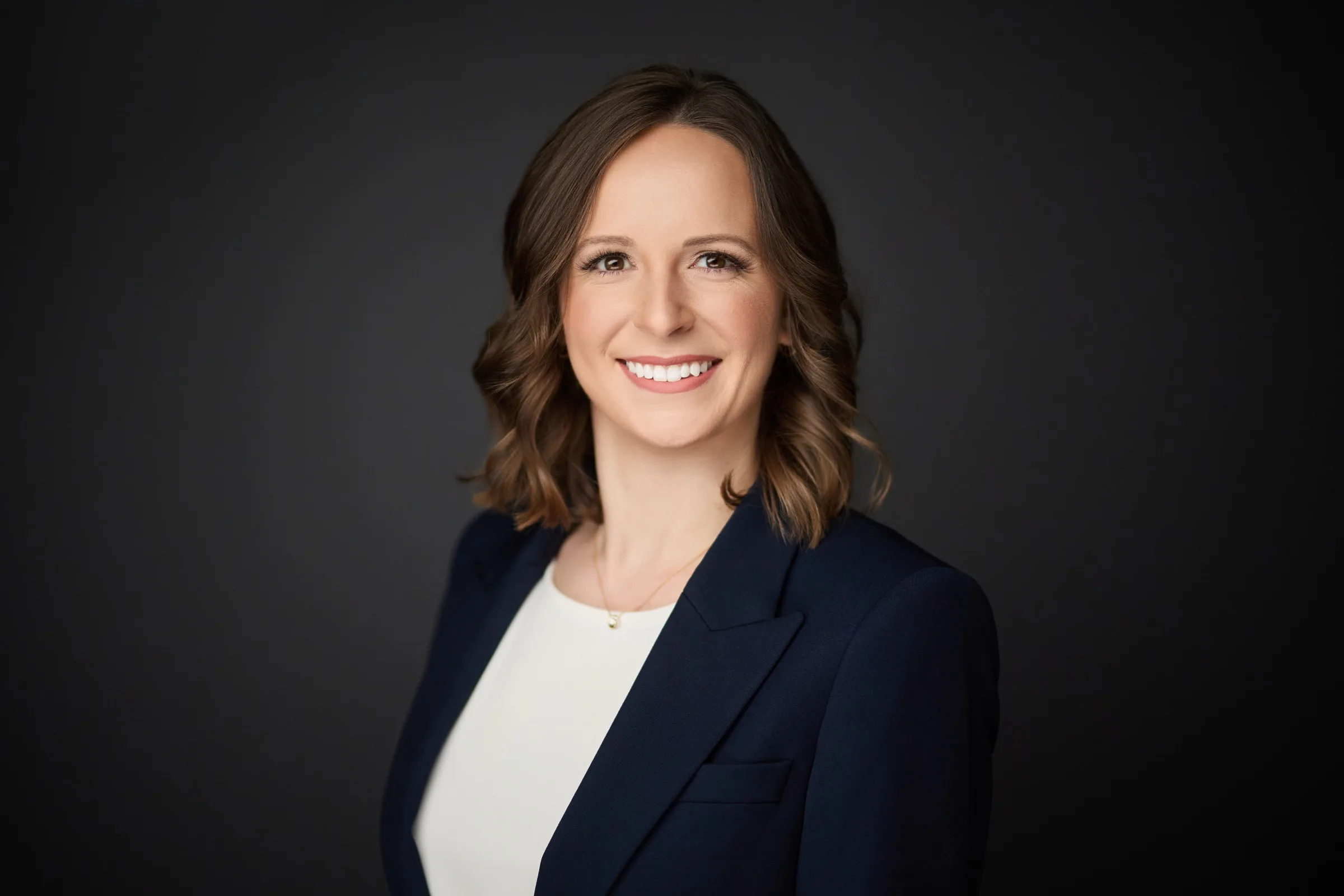 Emily, smiling to the camera in a navy blazer against a dark studio background.
