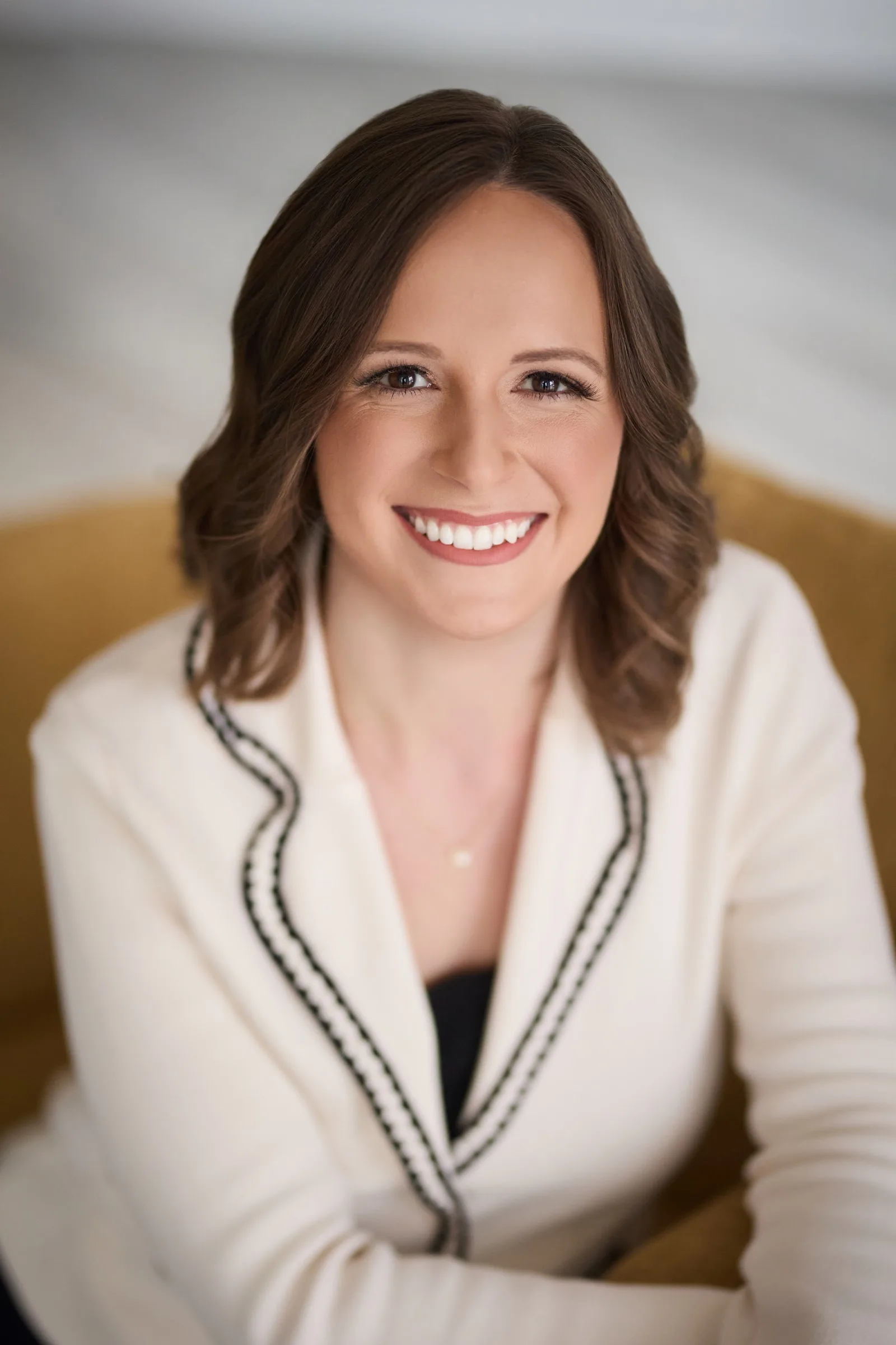 Emily, smiling to the camera in a cream jacket, warm studio light.